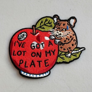 May include: Embroidered patch featuring a red apple with the text "I'VE GOT A LOT ON MY PLATE." A brown and tan mouse with a tear is perched on a green leaf next to the apple. The patch has a black border.