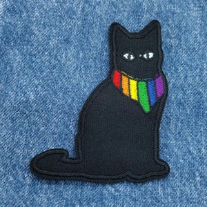 May include: Black cat with a rainbow bandana. The cat is sitting on a blue denim background.