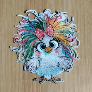 May include: Embroidered patch featuring a whimsical bird with large blue eyes, a red polka-dot bow, and colorful, feathery hair. The bird has a white body with blue and pink accents, and is standing on two orange feet. The patch is on a wooden surface.