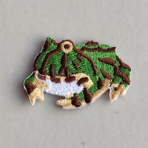 May include: Embroidered patch featuring a frog design. The patch has a green and brown camouflage pattern with white and tan accents. The frog patch is a unique accessory for clothing or bags.