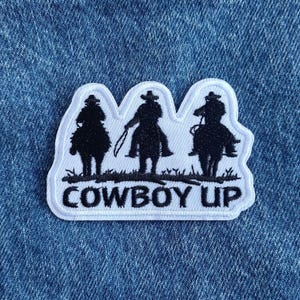 May include: White embroidered patch with three black silhouettes of cowboys riding horses. The text "COWBOY UP" is below the cowboys.