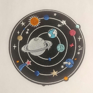 May include: Embroidered patch featuring a celestial design with a black background. The patch showcases a planet with rings, planets, stars, and a sun. The design includes various colors such as blue, orange, pink, and gold.