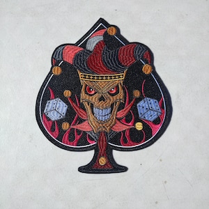 May include: Embroidered patch in the shape of a spade, featuring a skull wearing a jester's hat and a crown. The skull has red eyes and a wide grin. Flames and dice are also included in the design. The patch is black, red, gold, and blue.