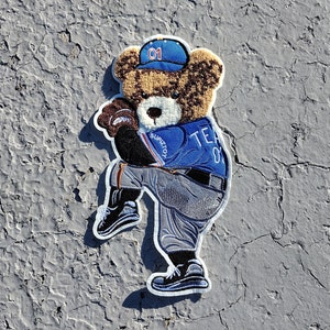 May include: A brown teddy bear wearing a blue baseball uniform with the number "01" on the hat and the text "TEAM 01" on the jersey. The bear is holding a baseball glove and is in a pitching position.