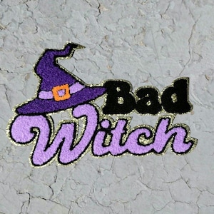 May include: A fabric patch featuring a purple witch hat with an orange buckle and the text Bad Witch in black and light purple lettering. The entire design is outlined with a shimmering gold border, creating a textured and decorative appearance.