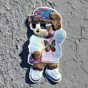 May include: A brown teddy bear wearing a rainbow tie-dye sweatshirt, a purple purse, sunglasses, and a blue and purple hat with a butterfly design. The bear is standing on a gray surface.