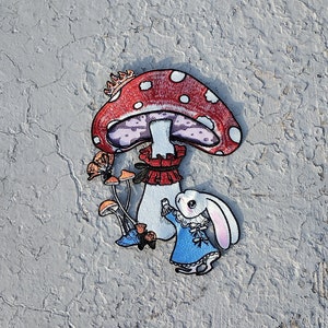 May include: A white rabbit wearing a blue dress with white lace trim stands next to a red and white mushroom with a gold crown. The mushroom has a purple underside and a red and white ruffle around the stem.