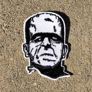 May include: Black and white embroidered patch of Frankenstein's monster's face. The patch is on a gray concrete background.