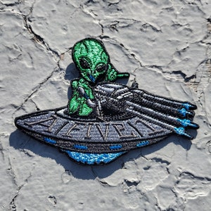 May include: Green alien with large eyes and a wide mouth sitting in a gray and blue UFO with the word "ALIEN" written on the side.