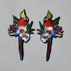 May include: Two embroidered iron-on patches featuring colorful parrots perched on tropical flowers and leaves. The parrots are red, yellow, blue, and green. The flowers are white, blue, and purple.