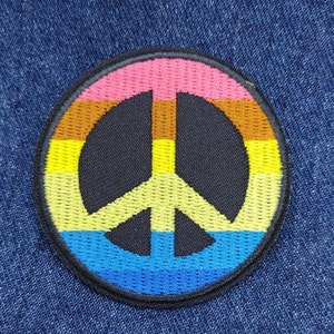 May include: A peace sign patch with a rainbow design. The patch is embroidered with yellow, orange, pink, blue, and black thread. The peace sign is a symbol of peace and love.