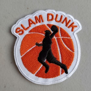 May include: Embroidered patch featuring a basketball design. The patch has a white border with the words "SLAM DUNK" in orange. Inside, a black silhouette of a basketball player is shown dunking a basketball over an orange basketball.