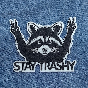 May include: Black and white embroidered patch of a raccoon giving the peace sign with the text "STAY TRASHY" below.