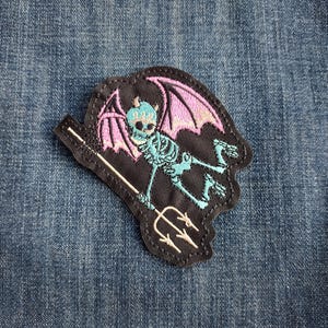 May include: Embroidered patch featuring a teal skeleton with pink wings, holding a trident. The patch has a black border and is set against a blue denim background. The skeleton has horns and is in a flying pose.