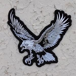 May include: A black and white embroidered patch of a bald eagle with its wings spread wide. The eagle is in flight and has a fierce expression on its face.