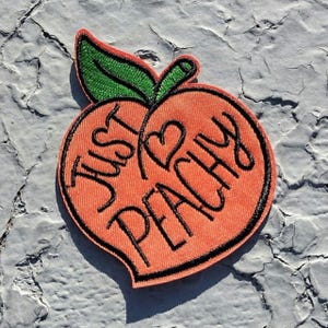 May include: Embroidered peach patch with the words "Just Peachy" in black script. The peach is orange with a black outline and a green leaf. A heart shape is included in the design.