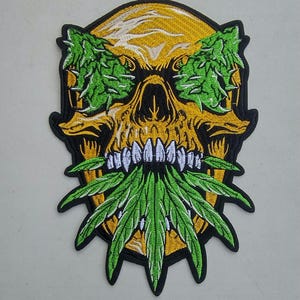 May include: A yellow skull with green marijuana leaves covering its eyes and mouth. The skull is embroidered on a black patch.