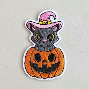 May include: Embroidered patch featuring a black cat wearing a pink witch hat, sitting inside an orange jack-o'-lantern. The patch has a white border and is designed for Halloween. The cat has yellow eyes and a pink nose.
