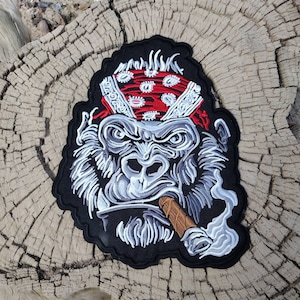 May include: Embroidered patch of a gorilla wearing a red bandana and smoking a cigar. The gorilla has a serious expression and is looking directly at the viewer.