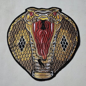 May include: A close-up of an embroidered patch featuring a cobra with its mouth open, showing its fangs. The cobra is gold and brown with red eyes and a red tongue. The patch is on a white background.