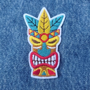 May include: Embroidered patch featuring a colorful tiki design. The patch has a white border and showcases a tiki mask with a yellow and brown leaf crown, blue leaves, and a red mouth with white teeth. The tiki's face is blue and brown.