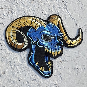 May include: A blue and gold embroidered patch of a skull with horns. The skull is open-mouthed with sharp teeth and has a menacing expression.