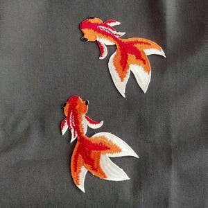May include: Two embroidered goldfish patches. The fish have red, orange, and white details. The patches are on a gray background. The fish are facing different directions.