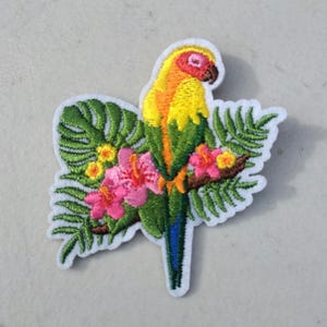 May include: Embroidered patch featuring a colorful parrot perched on a branch with pink flowers and green leaves. The parrot has yellow, orange, red, and green plumage. The patch has a white border and is likely for clothing or accessories.