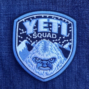 May include: A blue and white embroidered patch featuring a Yeti design. The patch has a shield shape with the words "YETI SQUAD" and a mountain range. The Yeti has a stern expression.
