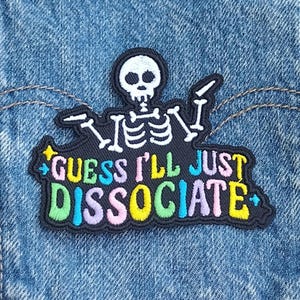 May include: Embroidered patch featuring a white skeleton with the text "GUESS I'LL JUST DISSOCIATE" in colorful letters. The patch is black with a colorful border and is set against a blue denim background.