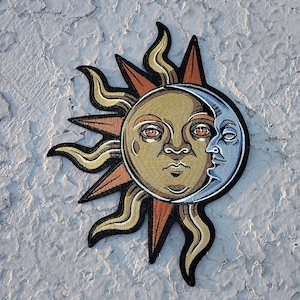 May include: A gold and blue embroidered patch depicting a sun and moon with faces. The sun has orange rays and the moon is a crescent shape.