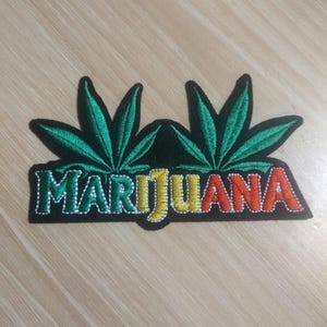 May include: Embroidered patch featuring two green cannabis leaves above the word "MARIJUANA" in green, yellow, and red lettering. The patch has a black background and white stitching.