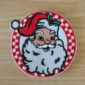 May include: Embroidered patch featuring Santa Claus's face. Santa is depicted with a white beard, rosy cheeks, and a red hat. The patch is circular, with a red and white checkered border.