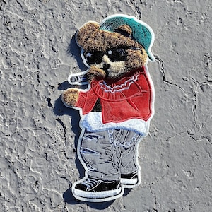 May include: Embroidered patch featuring a teddy bear wearing sunglasses, a red jacket, and a backwards baseball cap. The bear is holding a microphone and wearing gray jeans and black sneakers. The patch has a white border.