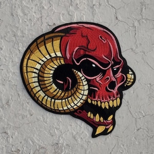 May include: Red and gold embroidered patch of a skull with horns. The skull has a fierce expression with sharp teeth and black eyes.