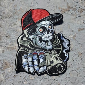 May include: A black and white embroidered patch of a skeleton wearing a red and black baseball cap and holding a gun. The skeleton has red eyes and a toothy grin.