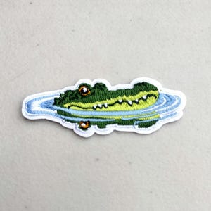 May include: Embroidered patch featuring a cartoon alligator in water. The alligator is green with yellow accents, and the water is blue. The patch is white around the edges.