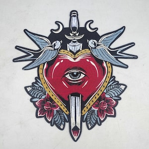 May include: Embroidered patch featuring a red heart pierced by a dagger, with an eye in the center. Two blue swallows flank the heart, with floral and leaf accents. The patch has a black background and gold trim.