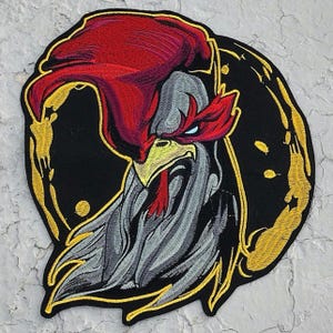 May include: An embroidered patch featuring a stylized rooster head with a prominent red comb, a yellow beak, and gray plumage. The design is set against a black circular background with a gold border and abstract gold accents surrounding the rooster.