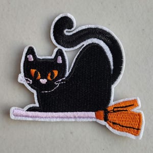 May include: Embroidered patch featuring a black cat riding a broom. The cat has orange eyes and white whiskers. The broom is orange with a pink handle, and the patch has a white border. A Halloween-themed item.