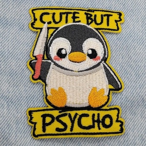 May include: An embroidered patch featuring a cartoon penguin with a black and white body, orange beak and feet, and pink cheeks. The penguin holds a small knife. Above and below the penguin are yellow banners with the black text CUTE BUT PSYCHO.