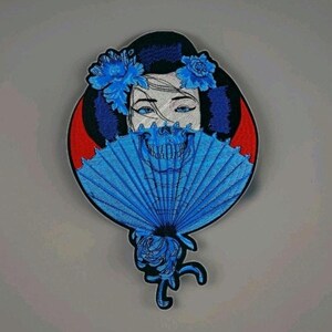 May include: A circular embroidered patch featuring a person with dark hair and blue flowers, holding a bright blue folding fan that obscures the lower face. The fan displays a skeletal jaw design, set against a red and black background.