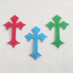 May include: Three colorful cross-shaped appliques in red, blue, and green. Each cross has a decorative design with pointed ends. The appliques are made of a felt-like material and are set against a white background.