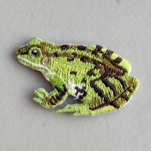 May include: An embroidered iron-on patch shaped like a frog. The frog is depicted in profile, featuring a vibrant lime green body with dark brown mottled spots across its back and legs. It has a white underbelly and a detailed black eye.