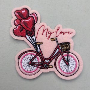 May include: Embroidered patch featuring a red bicycle with a basket and heart-shaped balloons. The text "My love" is stitched in red cursive above the bicycle. The patch has a pink background and white accents.