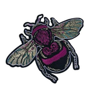 May include: An embroidered patch in the shape of a bumblebee. The bee features a black body with vibrant magenta pink stripes and intricate floral patterns on the thorax. The wings are detailed with metallic gold and teal thread stitching.