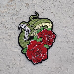 May include: Green snake with its mouth open, wrapped around red roses with green leaves. The snake and roses are embroidered on a black fabric.