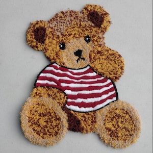 May include: A plush teddy bear applique with a red and white striped shirt. The bear is primarily brown and tan with dark brown accents on the ears and paws. The bear has black eyes and a black stitched nose.