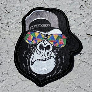 May include: Embroidered patch of a gorilla wearing a baseball cap and colorful sunglasses. The gorilla has a serious expression.