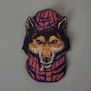 May include: An embroidered patch featuring a wolf wearing a pink and blue knit beanie and a matching scarf. The wolf has brown and black fur, pointed ears, and a cigarette in its mouth. The design uses detailed stitching to create texture.